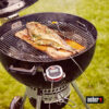 Ein ganzer Fisch wird auf einem Zedernholzbrett in einem schwarzen Weber Master-Touch Holzkohlegrill über einer grünen Rasenfläche gegrillt, ein digitales Thermometer zeigt die Temperatur an.