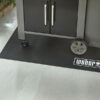 Schwarze Weber Bodenschutzmatte unter einem grauen Weber Gasgrill auf einer grauen Steinterrasse.