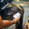Ein Mann hält ein Smartphone, das die Weber Connect App mit einer Temperaturanzeige von 95°C anzeigt, während ein großes Stück Fleisch auf einem Weber Holzkohlegrill gart.
