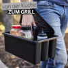 Eine Person in Jeans trägt einen schwarzen Weber Works Caddy mit Tragegriff und verschiedenen Gewürzen und Saucen. Der Text "von der Küche ZUM GRILL" ist über dem Caddy eingeblendet.