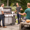 Drei Männer grillen und unterhalten sich im Freien auf einer gepflasterten Terrasse. Einer steht am offenen Weber Gasgrill, ein anderer schaut zu, und der dritte sitzt an einem Tisch mit Snacks.