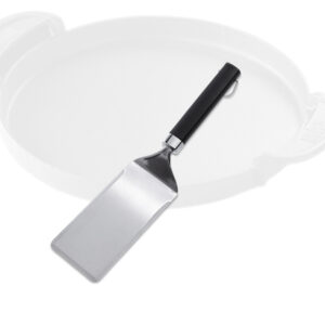Ein silberfarbener Weber Flat Top Durable Spatel aus Edelstahl mit einem schwarzen, rutschfesten Griff. Im Hintergrund ist der verschwommene Umriss einer hellen, runden Grillplatte mit zwei Griffen zu sehen.