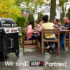 Eine Familie sitzt an einem Holztisch auf einer Terrasse und isst, während links ein schwarzer Weber Genesis II EX-335 Gasgrill steht. Im Vordergrund steht der Text "Wir sind: weber Partner!".