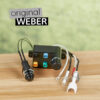 Schwarze Weber Zündbox mit Drehknopf, Zündelektrode und Kabeln auf einer hellbraunen Holzoberfläche vor einem verschwommenen grünen Hintergrund. Der Schriftzug "original WEBER" ist über dem Produkt platziert.