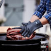 Eine Person trägt schwarze Grillhandschuhe, um ein mit Papier umwickeltes Lebensmittel auf einem Grill zu platzieren. Der Grill ist geschlossen, und im Hintergrund ist eine Person zu sehen.