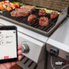 Ein Weber iGrill 3 Thermometer ist an einem Grill befestigt und misst die Temperatur von mehreren Steaks und Spießen. Ein Smartphone in einer Hand zeigt die zugehörige Weber iGrill App mit den aktuellen Temperaturen an.