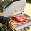 Eine Hand in einem schwarzen Grillhandschuh führt ein gelbes Weber iGrill Thermometer in ein großes rohes Steak auf einem Holzbrett neben einem Weber Spirit II Grill ein.