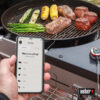 Ein Weber iGrill Mini Thermometer ist an einem Grill befestigt und misst die Temperatur eines Fleischstücks. Eine Hand hält ein Smartphone, das die Weber iGrill App mit der Auswahlmöglichkeit der Fleischsorte anzeigt. Auf dem Grill liegen außerdem Zucchini, Mais und Spargel.