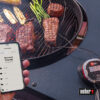 Ein Weber iGrill 2 Thermometer und ein Smartphone mit der Weber iGrill App neben einem Grill mit Steaks, Spargel und Maiskolben in Bregenz, Vorarlberg, Österreich.