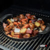 Ein schwarzer, runder Weber Original Gourmet BBQ System Einsatz, vermutlich eine Grillplatte oder eine gusseiserne Pfanne, gefüllt mit geröstetem Gemüse wie Kartoffeln, Karotten und Zwiebeln, auf einem Grillrost.