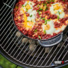 Ein schwarzer, runder Weber Gourmet BBQ System Einsatz, vermutlich eine gusseiserne Pfanne, gefüllt mit Shakshouka, verziert mit grünen Zwiebeln, auf einem Grillrost über Holzkohle.