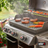 Ein silberfarbener Weber Genesis Gasgrill mit geöffnetem Deckel, auf dem Steaks, Karotten und Pfirsiche gegrillt werden. Rauch steigt von den Steaks auf.