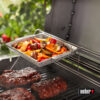 Ein silberner Weber Gemüsekorb aus Edelstahl mit bunten Paprika, Zwiebeln und Champignons, der auf einem Grillrost über glasierten Rippchen und Steaks platziert ist.